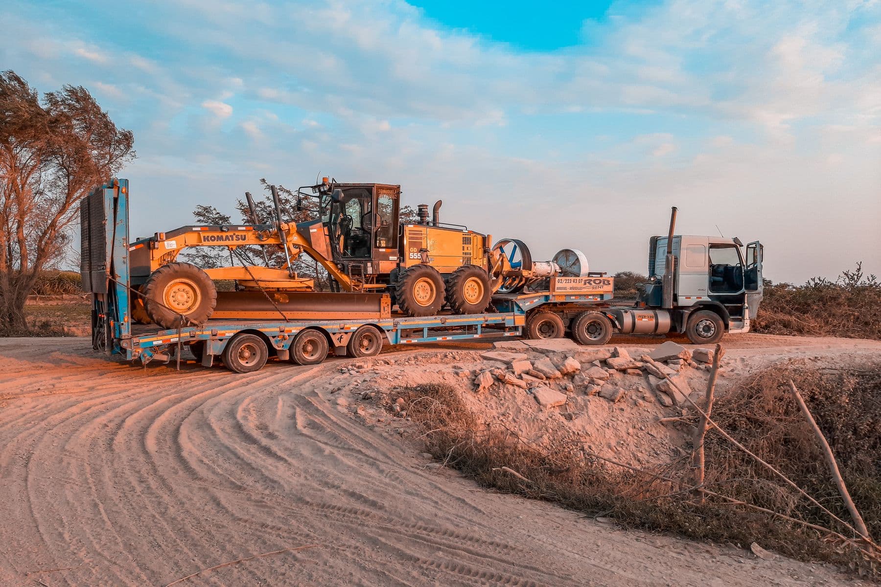 A cargo truck transporting heavy machinery on a rugged dirt road under a clear sky.