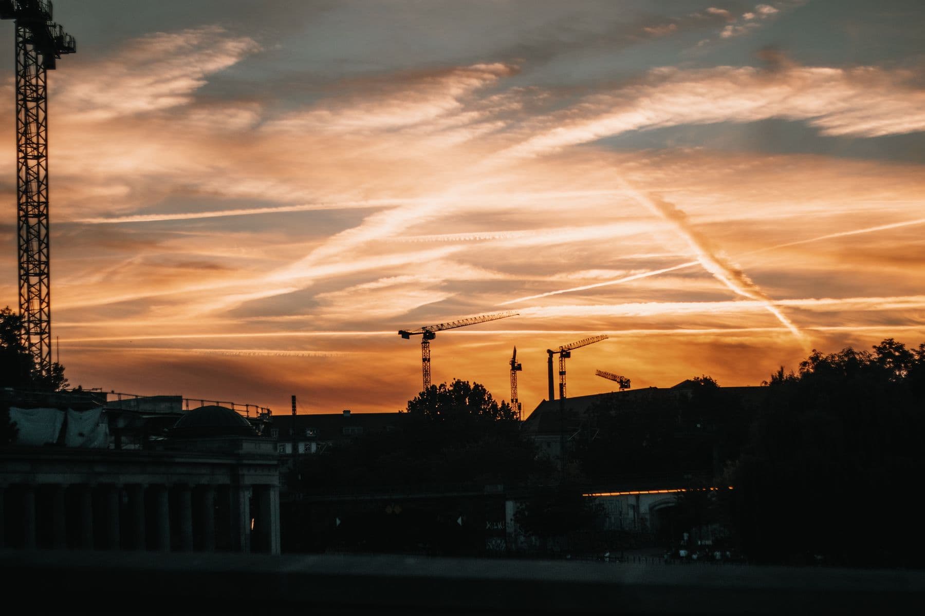 Silhouette of tower cranes against a dramatic sunset sky with orange hues and wispy clouds.